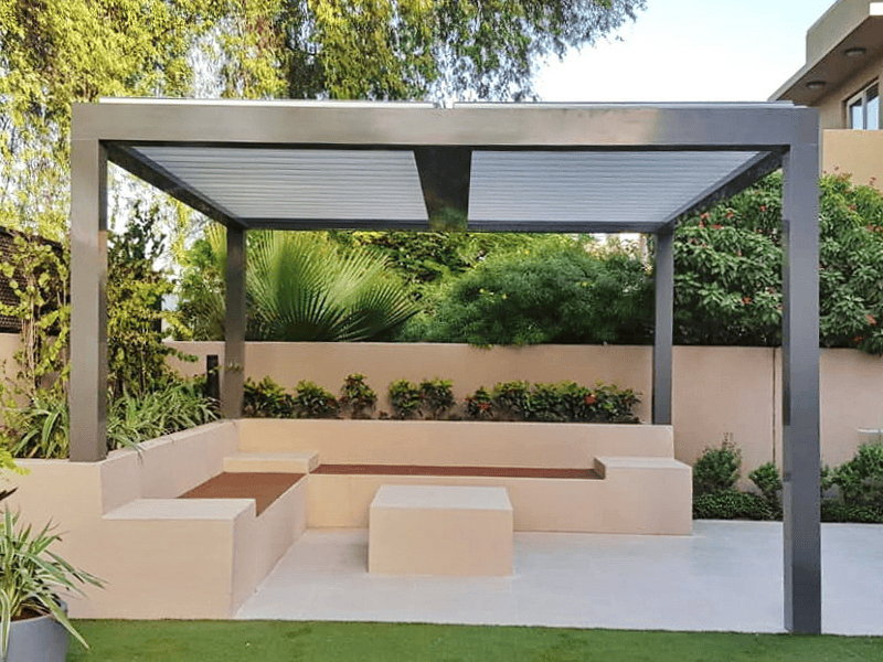 A modern outdoor patio in NY features a sleek pergola, built-in beige seating with wooden accents, a square coffee table, and lush green plants&mdash;a perfect inspiration for anyone seeking a sunroom contractor Long Island recommends.