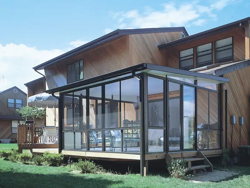 A modern NY house with wood siding features a glass-enclosed sunroom by a top sunroom contractor Long Island, set on a raised deck and surrounded by green grass and landscaping on a clear, sunny day.