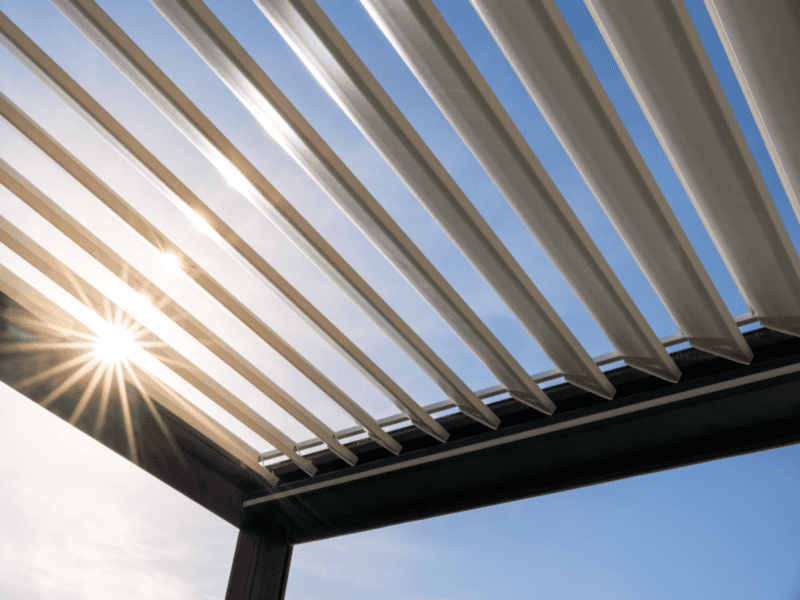 Sunlight shines through the slats of a modern pergola roof, creating shadows and lens flare against a clear blue sky, showcasing the craftsmanship of a sunroom contractor Long Island, NY homeowners trust.