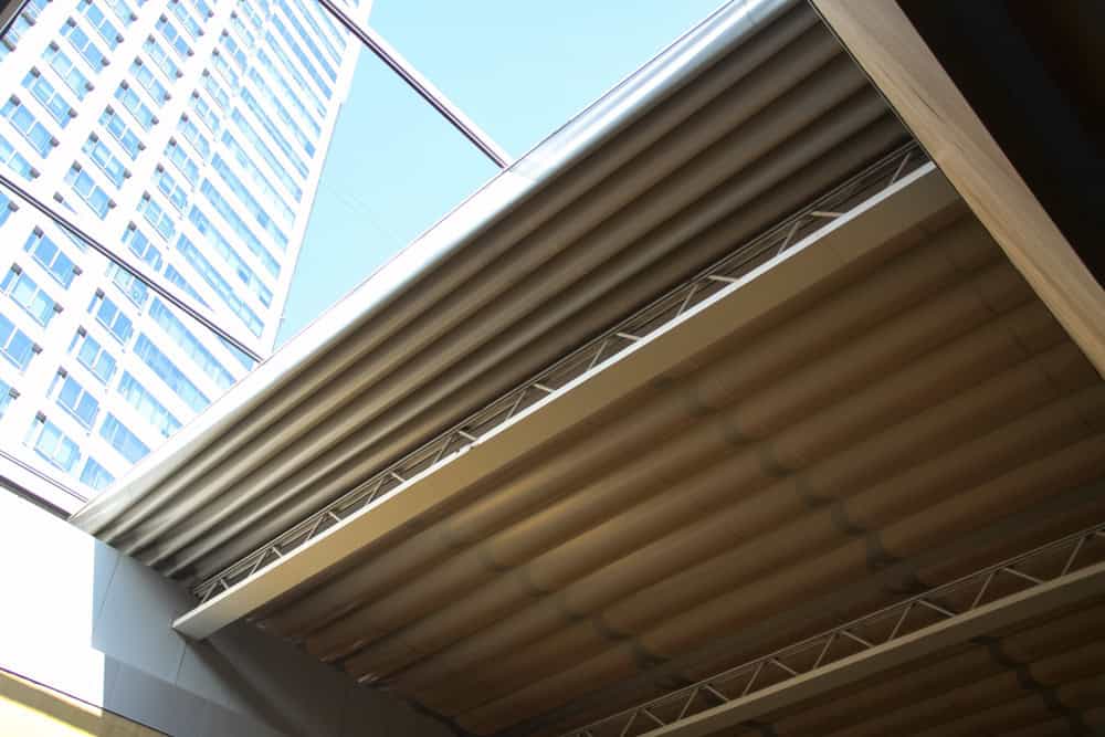 View from below of a partially open retractable roof with large metal panels&mdash;perfect inspiration for modern sunroom installation in Suffolk or Long Island&mdash;framed by a tall building and blue sky in the background.