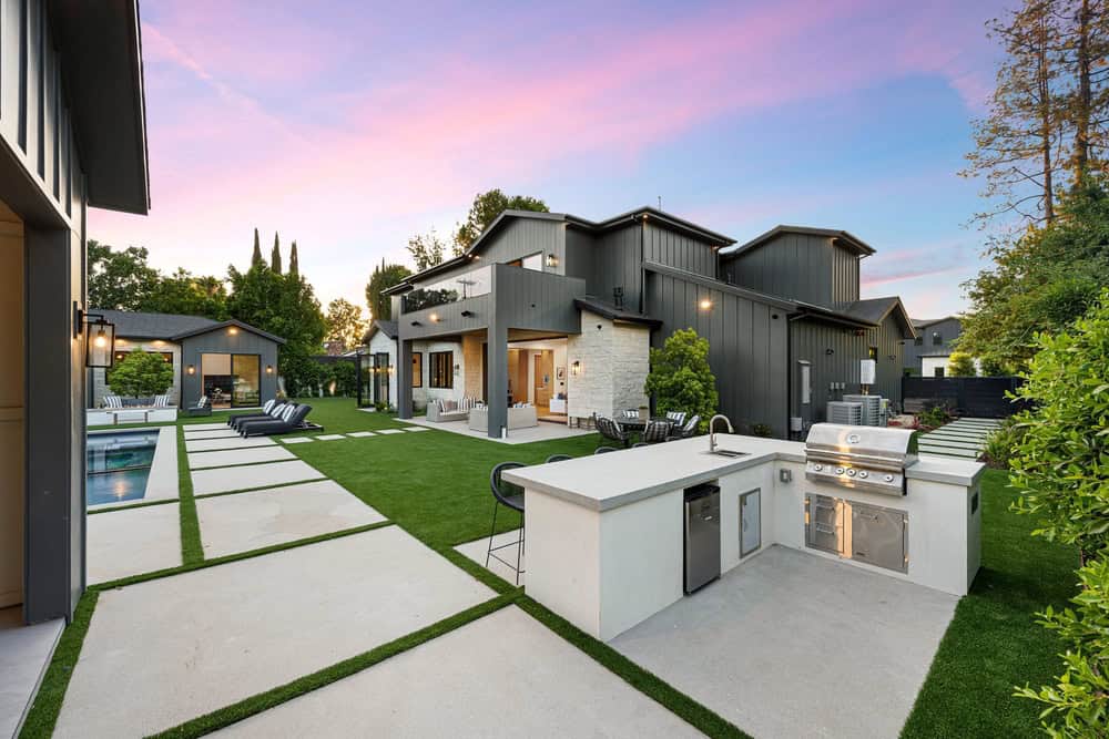 Modern backyard at sunset with a sleek gray house, all season sunrooms, outdoor kitchen with grill, patio seating area, pool, lounge chairs, green lawn, and concrete pavers creating geometric walkways&mdash;perfect for Suffolk or Nassau living.