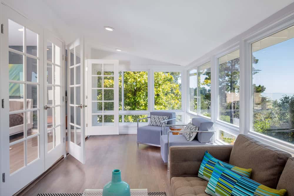 Bright sunroom with large windows, light wood floors, and white walls. Perfect for Long Island living, this Nassau sunroom installation features cozy gray armchairs, a brown sofa with colorful pillows, and views of sunlight and trees outside.