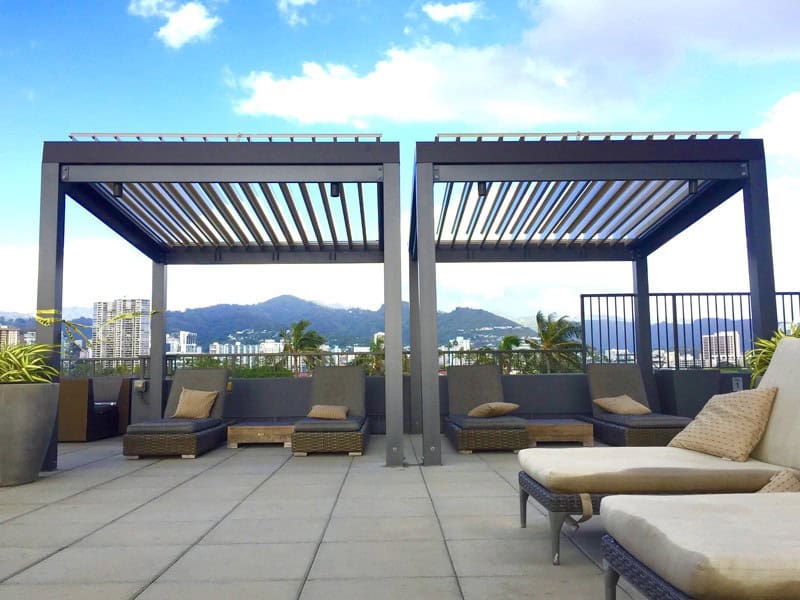 Modern rooftop lounge area with two pergolas, wicker lounge chairs, and potted plants&mdash;perfect for relaxing and enjoying city buildings and mountains. Inspired by stylish sunrooms Long Island, all under a partly cloudy NY sky.