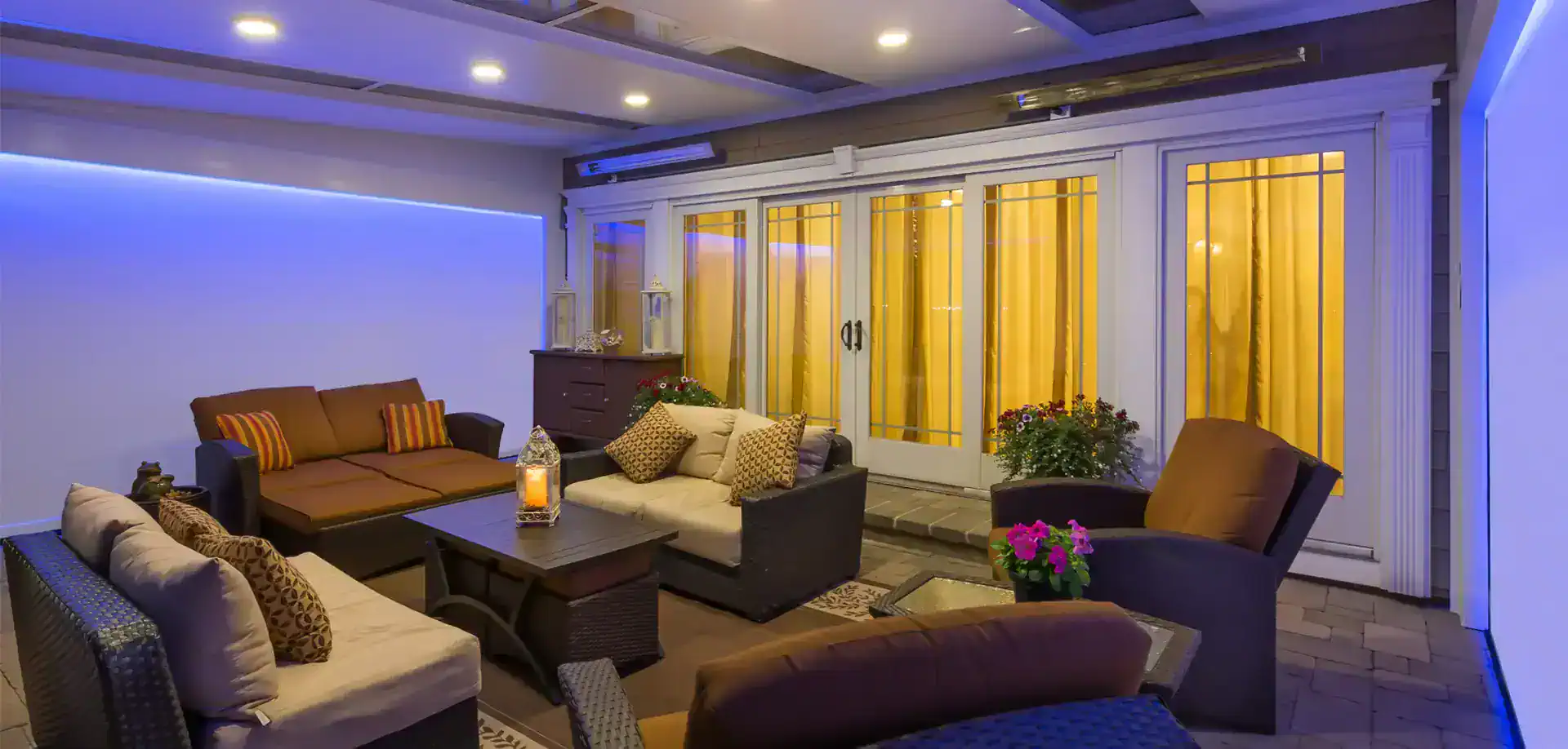 Outdoor patio with wicker furniture, beige and brown cushions, a central fire table, potted plants, and warm lighting. Sliding glass doors with vertical blinds lead inside. Blue accent lighting on the wall&mdash;perfect for TX evenings or custom sunrooms Houston style.