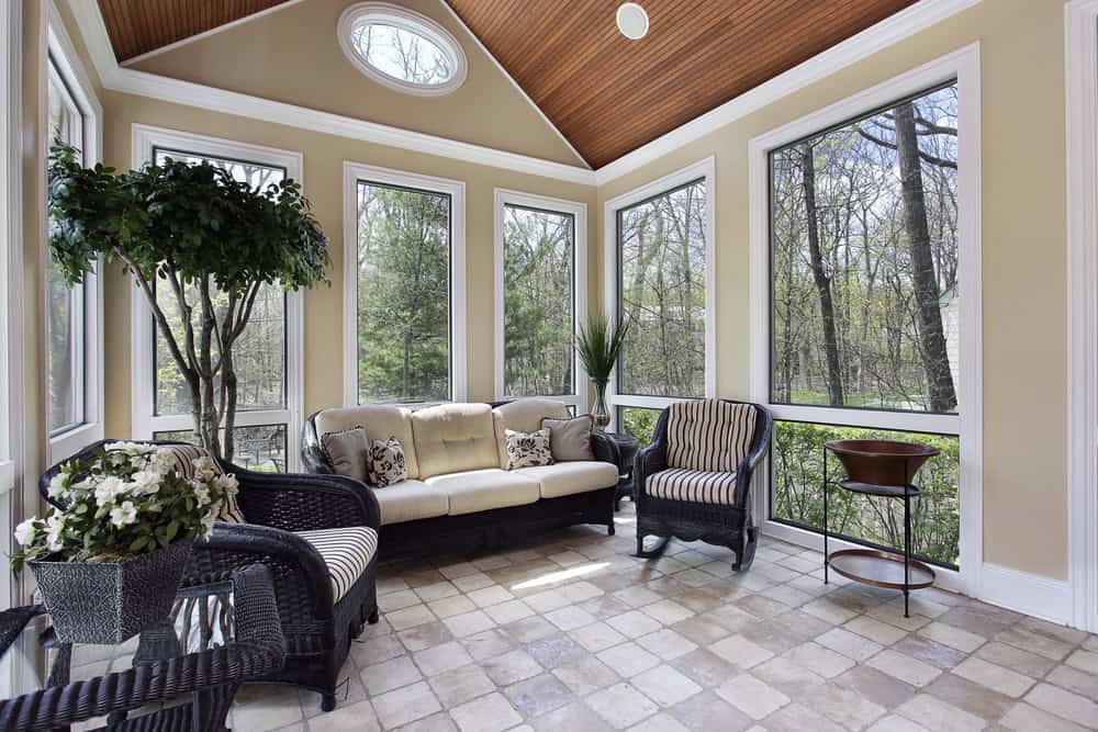 A bright sunroom with large windows, beige walls, tiled floor, black wicker furniture with cream cushions, potted plants, and a wood-paneled ceiling with a round skylight—perfect inspiration for custom sunrooms in Houston, TX.